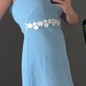 Ljusblå kort klänning från Jumper fabriken - Supersöt ljusblå kort klänning från Jumper med strukturmönster och vita blommiga detaljer i midjan. Klänningen är ärmlös och har rund halsringning samt dragkedja i ryggen. Perfekt för dig som gillar retrovibbar och vill sticka ut med en unik look.