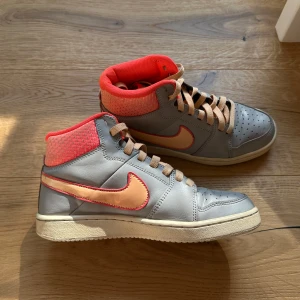 Unika Nike grå och korall höga sneakers - Unika Nike sneakers i high modell med grå skinnöverdel, korallfärgad mesh vid ankeln och beige snören. Swoosh-loggan är ljusorange med korallkant. Perforerad tå och platt sula ger en sportig vibe. Insidan är fodrad i stark korallfärg för extra kontrast.