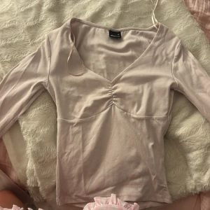 Ljusrosa långärmad topp Gina Tricot - Supersöt ljusrosa långärmad topp från Gina Tricot i storlek XS. Toppen har v-ringning, rynkdetalj framtill och är figurnära i modellen. Perfekt att styla med jeans eller kjol för en trendig look.
