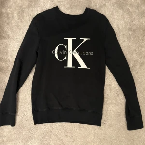 Svart sweatshirt från Calvin Klein - Svart sweatshirt från Calvin Klein Jeans med stor vit CK-logga framtill. Klassisk rund halsringning och långa ärmar. Tillverkad i mjuk bomullsmix som är skön att bära. Perfekt för en stilren och avslappnad look.