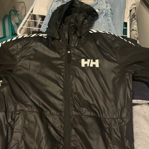 Svart vindjacka Helly Hansen med huva - Svart vindjacka från Helly Hansen med huva och hel dragkedja. Snygga vita ränder längs ärmarna och HH-logga på bröstet samt Helly Hansen-tryck på ryggen. Jackan är tillverkad i polyester och har sportig vibe.
