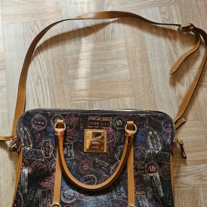 ALV datorväska med passstämpel-mönster - Snygg datorväska från ALV med passstämpel-mönster i flera färger på svart bakgrund. Väskan har bruna skinnhandtag, justerbar axelrem och guldfärgad logoplatta framtill. Invändigt finns flera fack och dragkedja. Perfekt för dig som vill sticka ut med din tech. Använd en gång och är i nyskick 