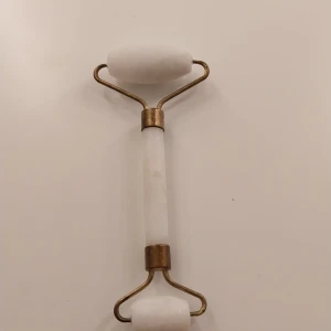 Vit ansiktsroller i sten - Säljer en vit ansiktsroller i sten med guldfärgade metalldetaljer. Rollern har två olika storlekar på stenarna, perfekt för både större och mindre partier i ansiktet. Materialet känns svalt och lyxigt mot huden.