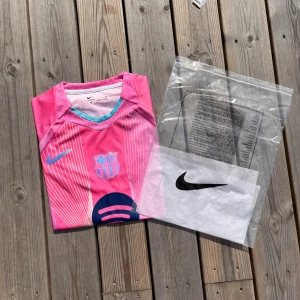 Barcelona Fotbollströja - Säljer en riktigt snygg Barcelona fotbollströja som är i helt nytt skick med etikett, förpackning och är helt oanvänd. Den är i storlek S och är i färgen rosa/vit med svarta detaljer. Pris:349kr men priset är inte hugget i sten. 