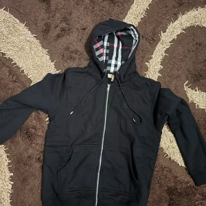 Svart hoodie från Burberry  - Svart hoodie från Burberry i storlek L med dragkedja och klassiskt rutigt foder i huvan. Tröjan har snörning vid huvan, två fickor framtill och en stilren look. Perfekt för dig som gillar exklusiva streetwear-vibbar.