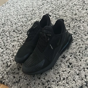 Svarta Nike Air Max 270 sneakers - Säljer ett par helsvarta Nike Air Max 270 sneakers med snygg mesh-överdel och stor synlig Air-enhet i hälen. Skorna har snörning och en sportig vibe, perfekta för dig som gillar streetstyle. Materialet är syntet och mesh för bra andningsförmåga.