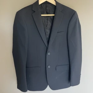 Svart kavaj Dressmann slim fit - Snygg svart kavaj från Dressmann i slim fit-modell. Klassisk design med två knappar framtill, fickor med lock och bröstficka. Tillverkad i ett slätt material med fodrad insida. Perfekt för dig som gillar stilrena och enkla plagg.