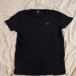 Svart t-shirt från Hollister - Svart t-shirt från Hollister med klassisk passform och liten broderad logga på bröstet. Tillverkad i 100% bomull, perfekt för en clean och enkel stil. Rund halsringning och korta ärmar gör den lätt att matcha med allt.