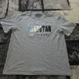 t-shirt - Snygg grå t-shirt från Trapstar med broderad logga i blått, vitt, grått och svart på bröstet. T-shirten har rund hals och korta ärmar, tillverkad i mjuk bomull. Perfekt för dig som gillar streetwear och vill sticka ut med coola detaljer. Storleken är S men jag skulle säga M.