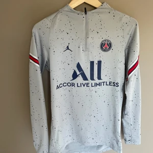 Grå långärmad PSG fotbollströja - Snygg grå långärmad PSG-tröja från Jordan med mörkblå logga och text, samt svarta prickar över hela tröjan. Röd, vit och blå rand på ärmarna ger extra stil. Materialet är lätt och andas, perfekt för träning eller match.