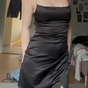 Svart satin klänning med smala axelband - Säljer en svart kort klänning i satin med smala axelband och figurnära passform. Klänningen har en slits framtill och en stilren, minimalistisk look som är perfekt för dig som gillar en chic vibe. Materialet är glansigt och mjukt mot huden.