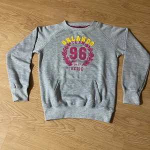 Grå sweatshirt från Oxide med tryck - Grå sweatshirt från Oxide med tryck i gult och rött framtill där det står 'Orlando Team 96'. Tröjan har rund hals, långa ärmar och en magficka. Mjuk insida och ribbade muddar vid ärmslut och nederkant. Perfekt för chill dagar.