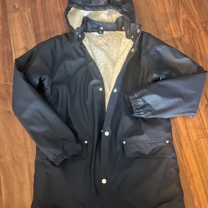 Mörkblå fodrad regnjacka Lager157 - Mörkblå regnjacka med vit teddyfoder från Lager157. Jackan har huva, tryckknappar framtill och två fickor med lock. Utsidan är i galon och insidan är mjuk och fluffig. 