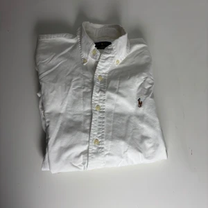 Vit skjorta från Ralph Lauren slim fit - Klassisk vit skjorta från Ralph Lauren i slim fit med broderad logga på bröstet. Skjortan har knappar hela vägen, button down-krage och är tillverkad i mjuk bomull. Perfekt för en clean och stilren look. Uppkom ett litet hål i tröjan varav priset går säkert att fixa 