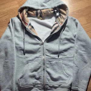 Grå hoodie från Burberry  - Snygg grå hoodie från Burberry med klassiskt rutigt foder i huvan. Tröjan har dragkedja framtill, snörning vid huvan och två fickor. Tillverkad i mjuk bomull för extra komfort. Perfekt för en chill och stilren look.