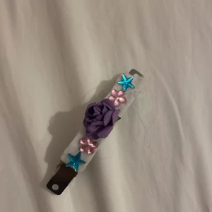 Hems jort hårspene med lila ros och stjärnor - Unikt hårsprne med vit bas, dekorerat med en lila tygros i mitten, två blå stjärnor och två rosa blommor i plast. Perfekt för dig som vill sticka ut med färgglada detaljer och blommor i håret.