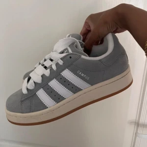 Gråa Adidas Campus sneakers i mocka - Snygga Adidas Campus sneakers i ljusgrå mocka med klassiska vita ränder och vit sula med gummidetalj. Skorna har breda vita snören och rund tå. Perfekta för dig som gillar en clean och stilren look.