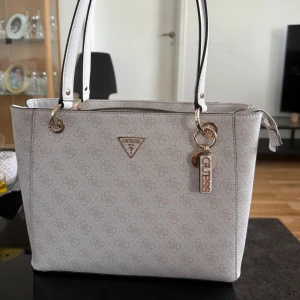 Guess väska  - Snygg vit handväska från Guess med diskret mönster och guldfärgade detaljer. Väskan har två långa handtag, dragkedja upptill och fina Guess-hängen. Perfekt storlek för att få plats med allt du behöver och ger en clean look till din outfit.