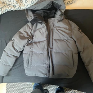 Grå pufferjacka från Canada Goose - Säljer en grå pufferjacka från Canada Goose med huva och snyggt broderad logga på ärmen. Jackan har dragkedja, stora fickor framtill och är quiltad för extra värme. Perfekt för kalla vinterdagar och har en clean, modern look.