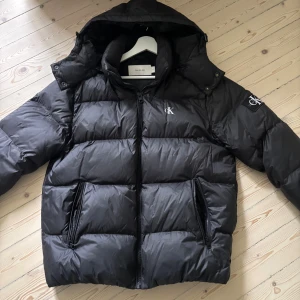 Svart pufferjacka Calvin Klein Jeans - Säljer en svart pufferjacka från Calvin Klein Jeans med huva och dragkedja framtill. Jackan har två stora fickor med dragkedja och logga på bröstet samt ärmen. Perfekt för kalla dagar nu när hösten och vintern börjar närma sig. //skick 9/10 nästan helt nu endast använd ett par fåtal gånger//nypris cirka 2500 mitt pris 1299, pris ej hugget i sten