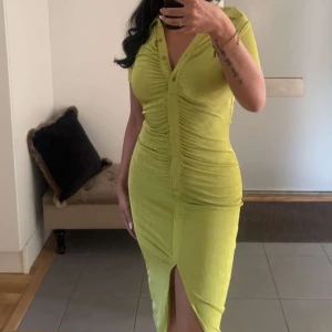 Grön figurnära klänning med slits - Snygg grön klänning med korta ärmar, v-ringning och knappar upptill. Klänningen är figurnära med rynkade detaljer längs magen och har en slits framtill. 
