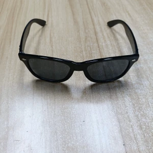 Svarta Ray-Ban Wayfarer solglasögon - Klassiska svarta Ray-Ban Wayfarer solglasögon med glansig båge och mörka glas. Snygg och tidlös design med Ray-Ban-logga på skalmen. Perfekta för att lyfta din stil under soliga dagar.