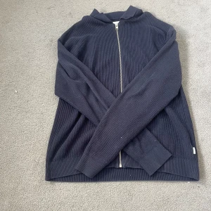 Mörkblå ribbad kofta med dragkedja - Säljer en mörkblå ribbad kofta med hel dragkedja framtill. Koftan har lång ärm och är tillverkad i ett stickat material som känns både mjukt och bekvämt. Perfekt för lager-på-lager och enkel att matcha med andra plagg. Storleken är Xs men den är hyfsat stor i storleken så passar även dig som har S