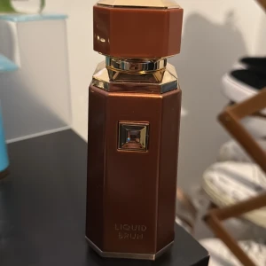 Liquid Brun parfymflaska brun/guld - Snygg parfymflaska från Liquid Brun i en lyxig brun färg med guldiga detaljer och geometrisk form. Flaskan har en elegant, fyrkantig design med en dekorativ sten på framsidan och texten 'LIQUID BRUN' ingraverad längst ner.