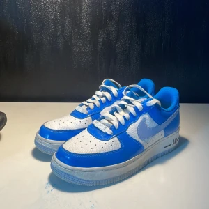Nike Air Force 1 blå och vita sneakers - Nike Air Force 1 sneakers i blått och vitt med klassisk låg siluett. Ovandel i skinn med perforerade detaljer och vit swoosh på sidorna. Tjock vit sula med AIR-detalj, vita skosnören och blå insida. Perfekt för dig som vill sticka ut med en färgstark look.