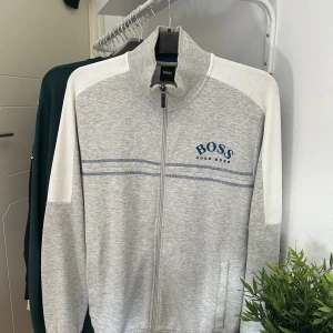 Grå ziptröja från Hugo Boss - En väldigt fin Hugo boss zip (sällsynt) i storlek L, men passar även M. Tröjan är i ljusgrå färg med blåa detaljer. Köptes när tröjan fortfarande såldes på marknaden. 