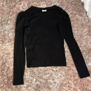 Svart långärmad tröja från JDY - En svart, långärmad tröja från JDY i storlek XS. Tröjan har en enkel, rund halsringning och är figurnära med lätt puffade axlar för en trendig touch. Perfekt basplagg att styla med jeans eller kjol.