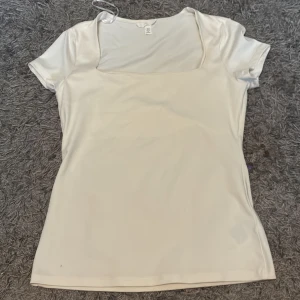 Vit topp med fyrkantig ringning H&M - En stilren vit topp från H&M med korta ärmar och snygg fyrkantig ringning. Toppens material har en lätt glansig finish som ger en fräsch look. Perfekt att matcha med jeans eller kjol för en clean vibe.