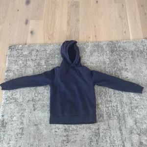 Mörkblå hoodie från These Glory Days - Mörkblå hoodie från These Glory Days i 100% ekologisk bomull. Klassisk modell med huva och snörning, samt långa ärmar och ribbade muddar. Perfekt för en chill och avslappnad stil.