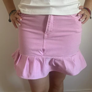 Rosa jeanskjol med volangkant - Supersöt rosa jeanskjol med volangkant längst ner. Kjolen har klassiska fickor både fram och bak samt en snygg söm framtill. Perfekt för dig som gillar färg och vill sticka ut lite extra. Passformen är normal och materialet är jeans.