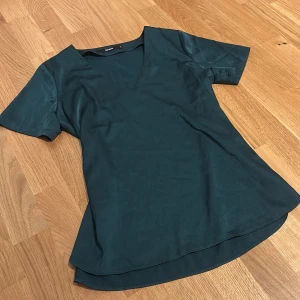 Mörkgrön blus från BikBok - Snygg mörkgrön blus från BikBok i slätt, glansigt material. Modellen har v-ringning, korta ärmar och lätt utställd nederkant. Perfekt för dig som gillar stilrena och enkla plagg med en elegant touch.
