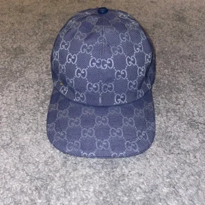 Blå keps från Gucci med GG-mönster - Snygg blå keps från Gucci med ikoniskt GG-mönster över hela kepsen. Kepsen har böjd skärm och en liten guldfärgad GG-detalj på sidan. Tillverkad i ett slitstarkt tyg med vävd struktur, perfekt för att lyfta din streetwear-look.