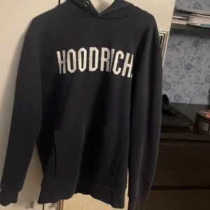 Svart hoodie från Hoodrich med tryck - Svart hoodie från Hoodrich i storlek small med vit HOODRICH-logga framtill. Klassisk modell med huva och känguruficka. Perfekt för dig som gillar streetwear och vill ha en stilren look.