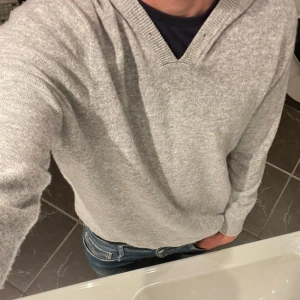 Grå stickad hoodie med v-ringning - Mysig grå stickad hoodie med v-ringning och huva. Tröjan har lång ärm och ribbade muddar vid ärmslut och nederkant. Perfekt för kyliga dagar och enkel att matcha med jeans eller andra byxor.