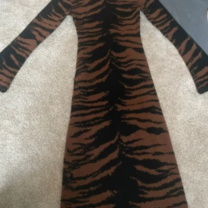 Stickad långklänning med zebramönster - Snygg lång stickad klänning med zebramönster i svart och brunt. Klänningen har lång ärm och figurnära passform som framhäver siluetten. Perfekt för dig som vill sticka ut med ett djurmönster och ändå hålla det bekvämt och trendigt.