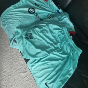 FC Barcelona Nike fotbollströja turkos - FC Barcelona matchtröja och shorts i turkos från Nike. Tröjan har korta ärmar, tryckta loggor från Spotify och UNHCR samt klubbmärke på bröstet. Materialet är lätt och andas, perfekt för fotboll. Diskret randigt mönster i rött och blått över hela tröjan.
