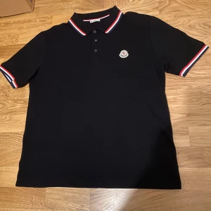 Svart Moncler pikétröja med röd/vita detaljer - Svart pikétröja från Moncler med klassisk krage och knäppning framtill. Tröjan har röda och vita ränder på krage och ärmslut samt Moncler-logga broderad på bröstet (NFC tagg). Perfekt för en clean och sportig look.