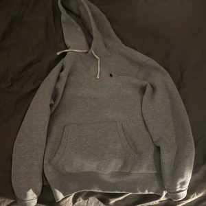 Söker Byte Från M till S, Grå Ralph Lauren Hoodie - Snygg grå hoodie från Polo Ralph Lauren med klassisk huva och vit snörning. Tröjan har en liten broderad logga på bröstet och en stor magficka framtill. Tillverkad i mjuk bomullsmix, perfekt för chill dagar.Äger i M men söker likadan i S. Denna är även bara använd få tal gånger.
