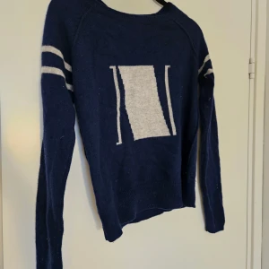 Mörkblå stickad tröja i kashmir  - Mörkblå stickad tröja i kashmir. med ljusgrå ränder på ärmarna och mönster framtill. Tröjan har rund halsringning och långa ärmar.