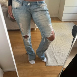 Ljusblå slitna bootcut jeans - Säljer ett par ljusblå jeans med slitningar och stora hål vid knäna. Jeansen har bootcut-modell och låg midja, med klassiska fem fickor och dragkedja. Perfekta för dig som gillar en avslappnad och edgy stil.