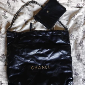 Svart quiltad axelväska  - Säljer en svart quiltad axelväska i mjukt skinn med silverfärgad kedjerem och tillhörande liten pouch med klassisk loggal. Perfekt för dig som vill ha en lyxig och tidlös accessoar. Materialet ska vara läder enligt säljaren där jag köpte men jag reserverar mig till att inte lova det på 100% då det är olika hur det uppfattas, jag nedöm det dock som läder. Ingår dustbag.