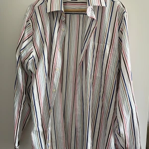 Randig skjorta från Paul Smith London - Snygg skjorta från Paul Smith London, tillverkad i Italien. Skjortan har ett färgglatt randigt mönster i vitt, blått, rött, rosa, brunt och grönt. Klassisk krage, långärmad och med knappar framtill samt manschetter. Perfekt för dig som gillar stilrena och unika plagg.