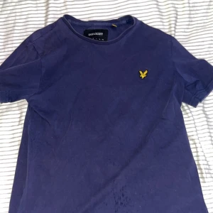 Marinblå t-shirt från Lyle & Scott - Snygg marinblå t-shirt från Lyle & Scott med klassisk rund hals och korta ärmar. På bröstet sitter den ikoniska gula örnloggan broderad. Tillverkad i mjuk bomull för en skön känsla hela dagen. Har använt den några gånger den har små små mini håll där nere på framsidan men dom syns knappast. 