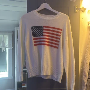 Vit stickad tröja med USA-flagga - Vit stickad tröja med ribbad rund hals och muddar. På bröstet finns en stor USA-flagga i blått, rött och vitt. Tröjan har lång ärm och en loose passform som ger en chill vibe. Perfekt för dig som gillar statement-plagg och vill sticka ut.