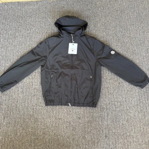Svart vindjacka från Moncler med huva - Storlek L passar M. Svart vindjacka från Moncler med huva, hel dragkedja och två fickor framtill med dragkedja. Jackan har elastiska muddar och midja samt Moncler-logga på ärmen. Tillverkad i polyester och har en clean, sportig look.