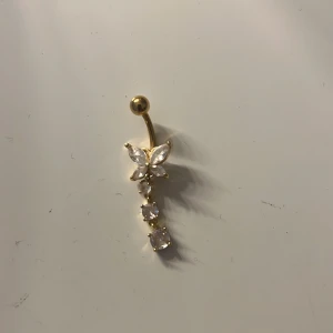 Guldig navelpiercing med fjäril - Guldfärgad navelpiercing barbell med en fjäril. Materialet i staven är kirurgiskt stål (316L). Staven är 10 mm lång och 1,6 mm bred. Den är köpt från ur& Penn 
